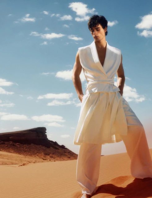 A fair-skinned man poses in a white sleeveless top and flowing white pants, set against a sandy desert landscape under a bright sky.