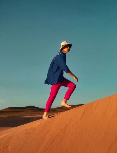Adult strides barefoot up a sand dune wearing a hat, blue shirt, and pink pants under a clear blue sky on a sunny day outdoors.