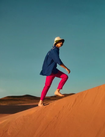 Adult strides barefoot up a sand dune wearing a hat, blue shirt, and pink pants under a clear blue sky on a sunny day outdoors.