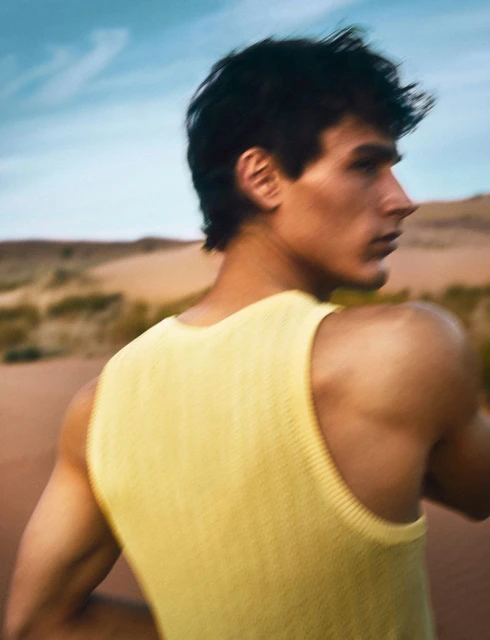 Portrait of an adult male wearing a yellow tank top posing in the desert under a blue sky. The landscape is dry and sandy.