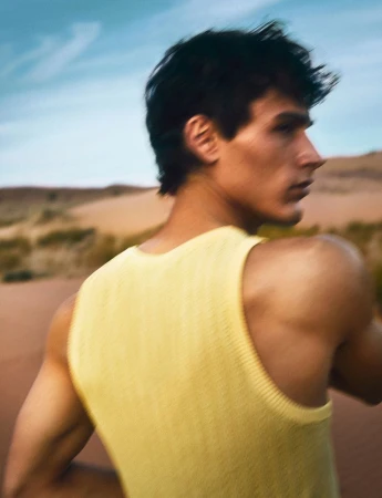 Portrait of an adult male wearing a yellow tank top posing in the desert under a blue sky. The landscape is dry and sandy.