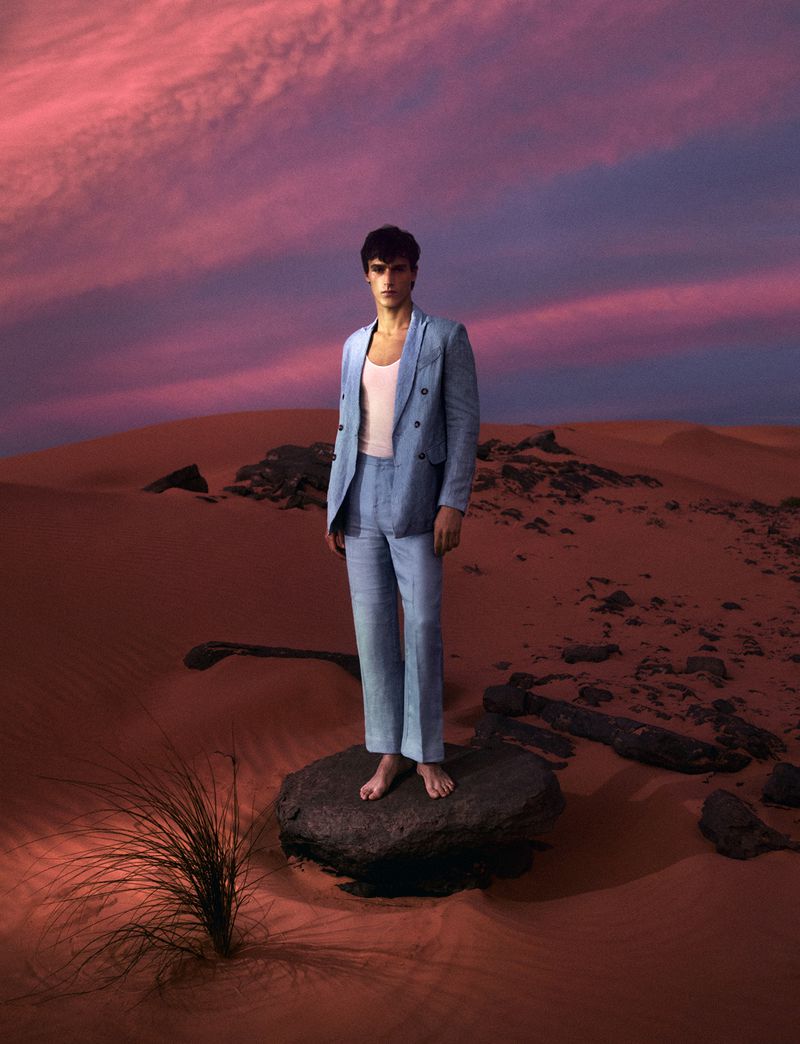 A stylish man in a light blue linen suit stands on a rock in the desert at dusk, against a vivid pink sky.