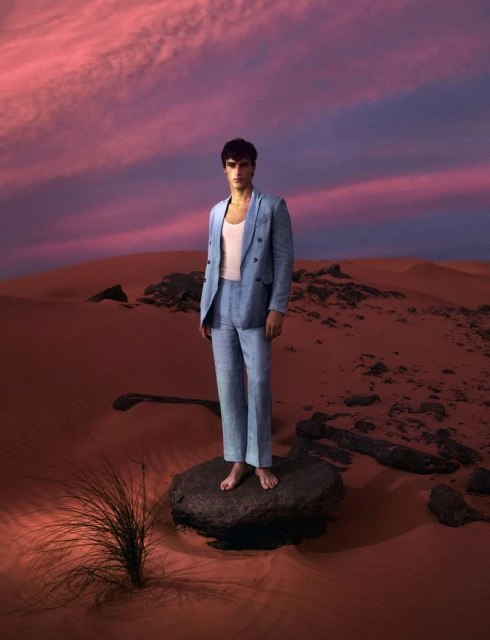 A stylish man in a light blue linen suit stands on a rock in the desert at dusk, against a vivid pink sky.