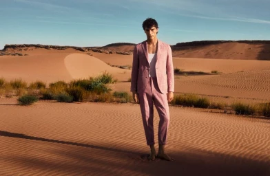 Male model standing barefoot in a desert landscape wearing a pink suit and white t-shirt under open daylight.