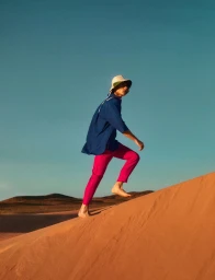 Model wearing a blue shirt and bright pink trousers walking barefoot up a sandy dune under a clear sky