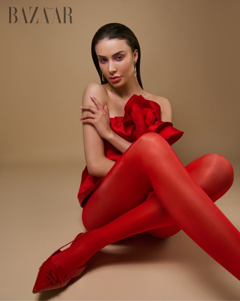 Elegant woman in red dress and stockings poses in a fashion shot against a neutral backdrop. She wears gold earrings.