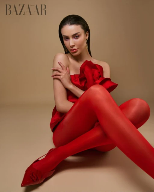 Elegant woman in red dress and stockings poses in a fashion shot against a neutral backdrop. She wears gold earrings.
