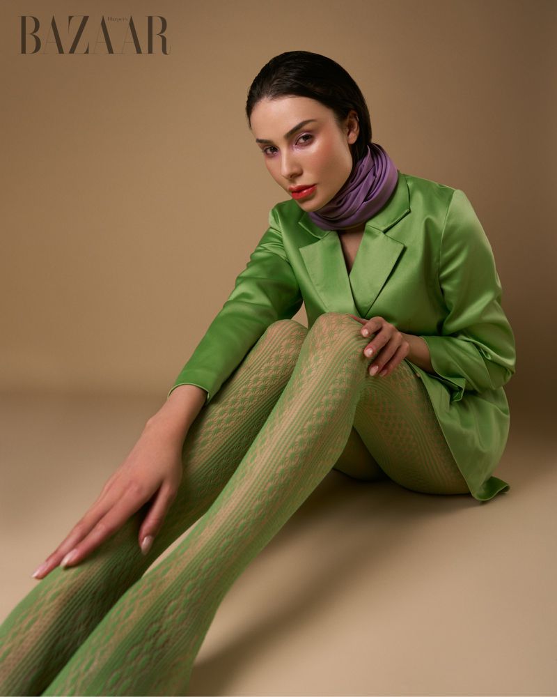 A young adult woman poses in a photo studio, wearing a green blazer, light-green textured tights, and a purple scarf, against a neutral backdrop.