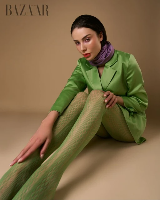 A young adult woman poses in a photo studio, wearing a green blazer, light-green textured tights, and a purple scarf, against a neutral backdrop.