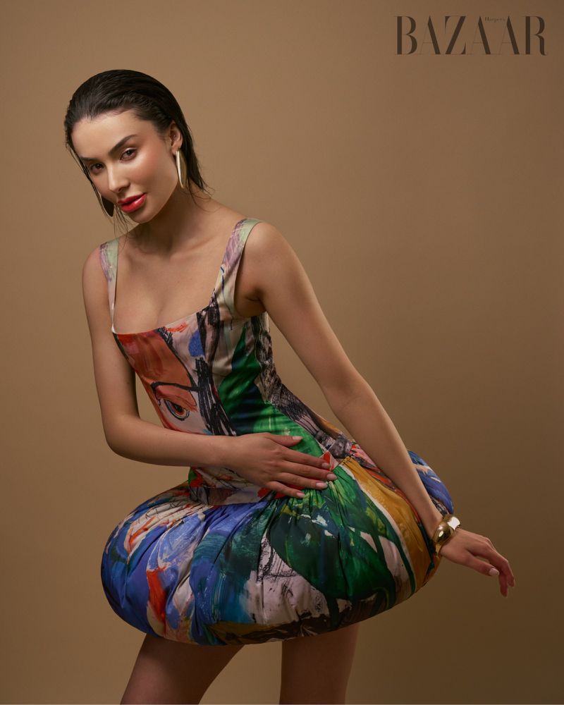 A woman poses in a colorful, short dress against a tan backdrop with Bazaar branding, showcasing a high-fashion editorial look.