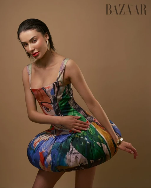 A woman poses in a colorful, short dress against a tan backdrop with Bazaar branding, showcasing a high-fashion editorial look.