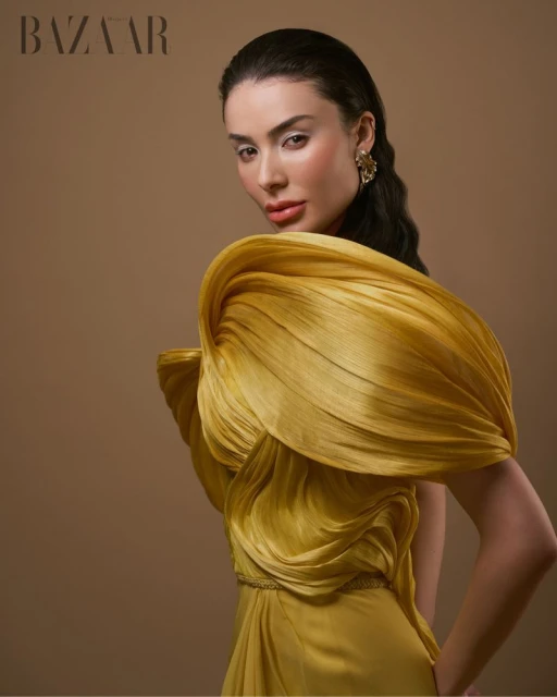 Elegant adult woman in a yellow couture dress posing in a studio for a fashion shot with the Bazaar logo visible in the background.