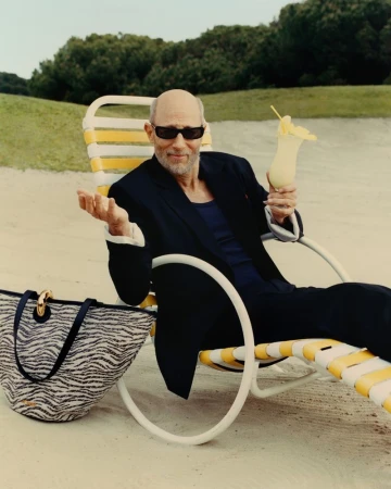 Jon Gries seated in a yellow-and-white lounge chair on a sandy beach holding a cocktail, with a patterned tote bag beside him.