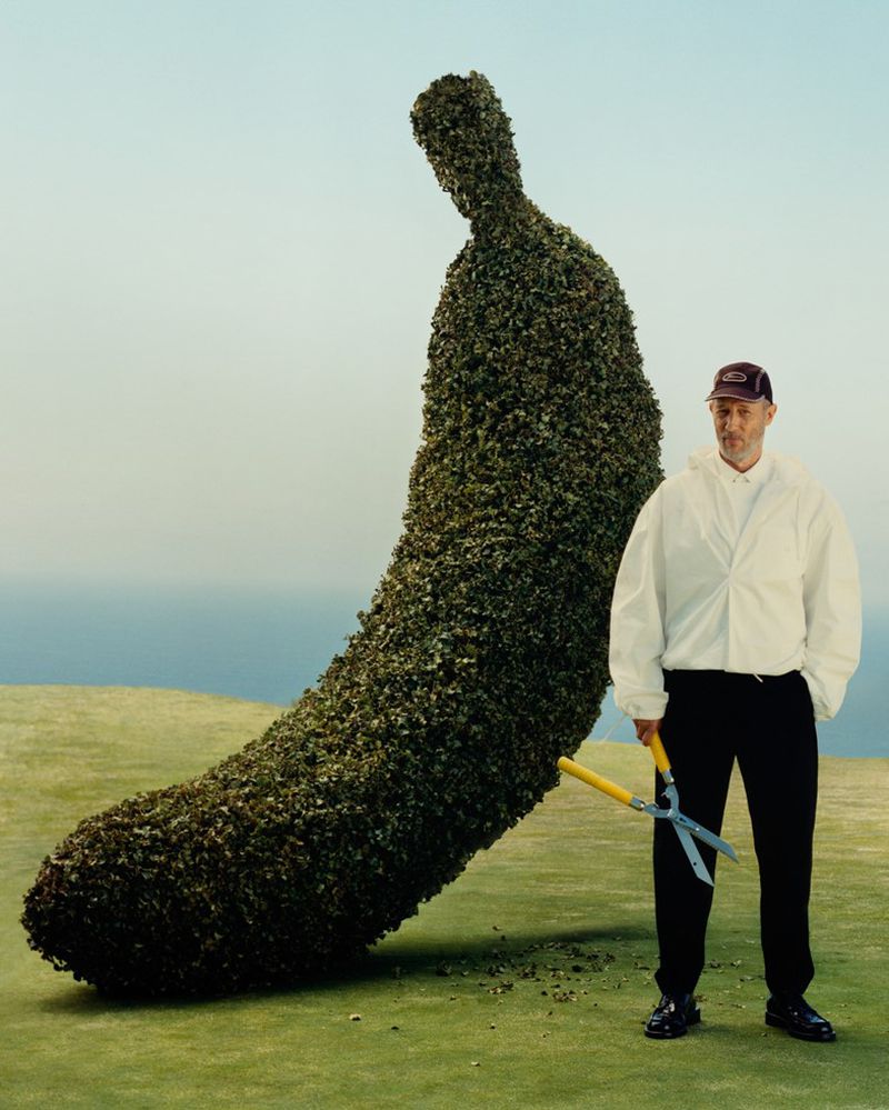 Adult male gardener stands with hedge shears beside a large banana-shaped topiary on a green lawn with a light blue sky backdrop.