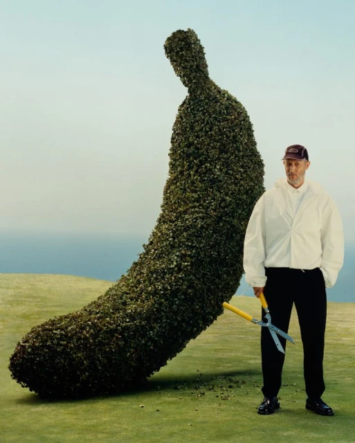 Adult male gardener stands with hedge shears beside a large banana-shaped topiary on a green lawn with a light blue sky backdrop.