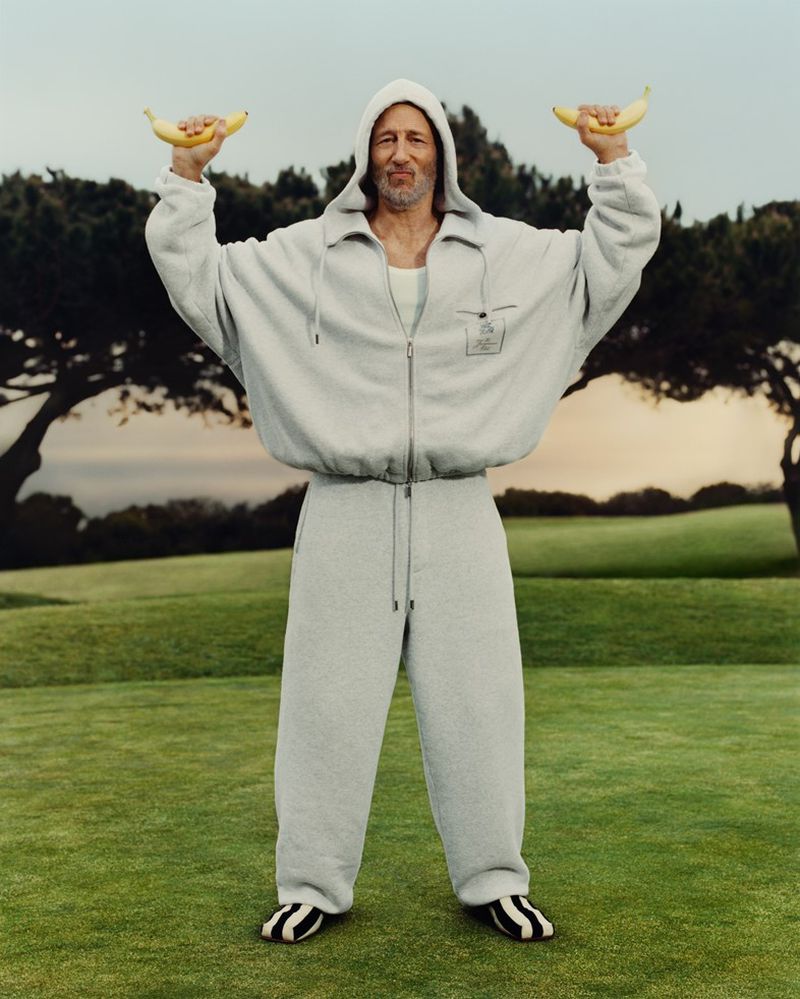 An adult male in a gray tracksuit stands on a green lawn, holding a banana in each hand with trees and a blue sky in the background.