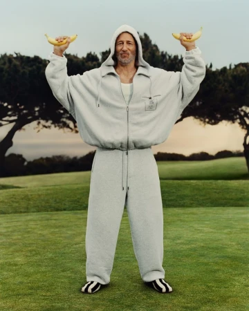 An adult male in a gray tracksuit stands on a green lawn, holding a banana in each hand with trees and a blue sky in the background.