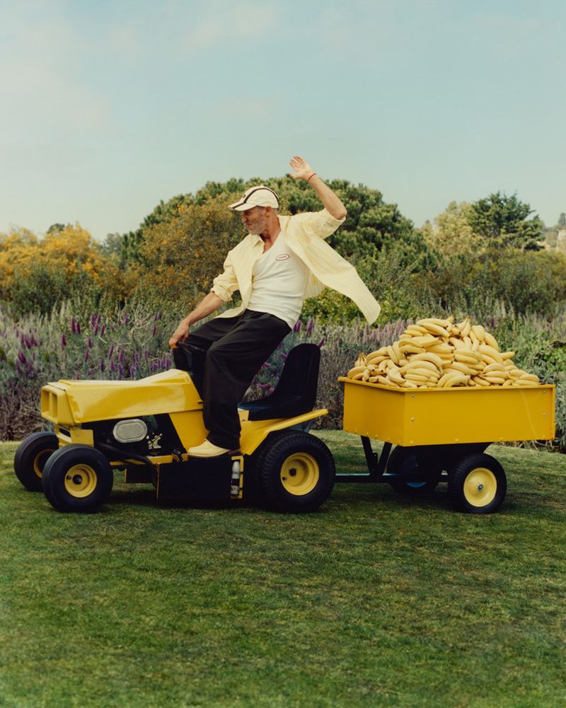 Man rides a yellow lawn mower pulling a banana-filled trailer on green grass, wearing a white shirt, cap, and dark pants, under a blue sky.