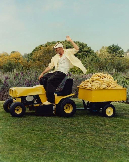 Man rides a yellow lawn mower pulling a banana-filled trailer on green grass, wearing a white shirt, cap, and dark pants, under a blue sky.