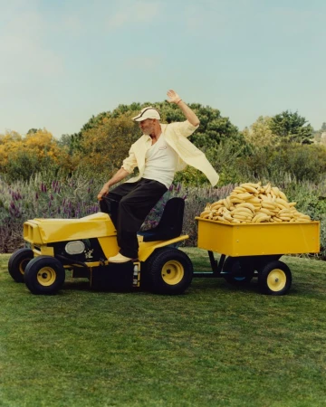 Man rides a yellow lawn mower pulling a banana-filled trailer on green grass, wearing a white shirt, cap, and dark pants, under a blue sky.