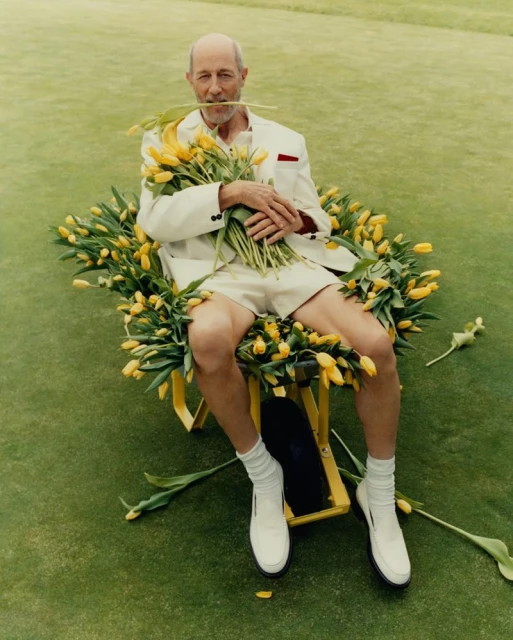 Elderly man in cream suit sits on a yellow chair amidst a field of yellow tulips, holding a bouquet, on a green lawn.