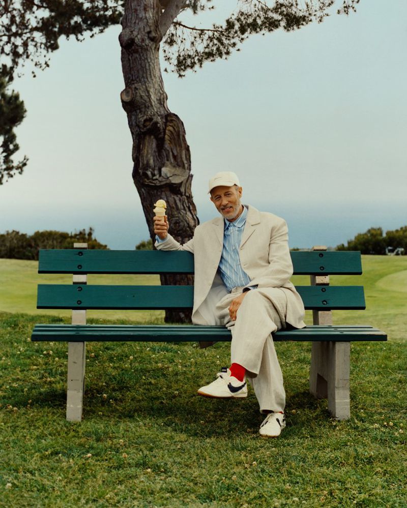 Mature man sits on a park bench outdoors in a cream suit, holding an ice cream cone on a sunny day with a golf course in the background.