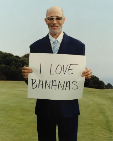 Jon Gries wearing a dark suit and sunglasses holding a handwritten sign saying I Love Bananas outdoors on a grassy lawn