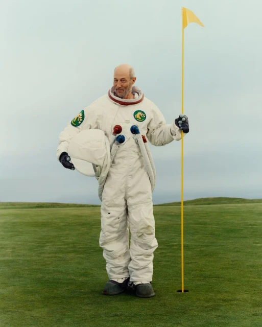 Mature man in astronaut suit poses next to golf hole on lush green course under overcast sky, holding helmet and golf flag.