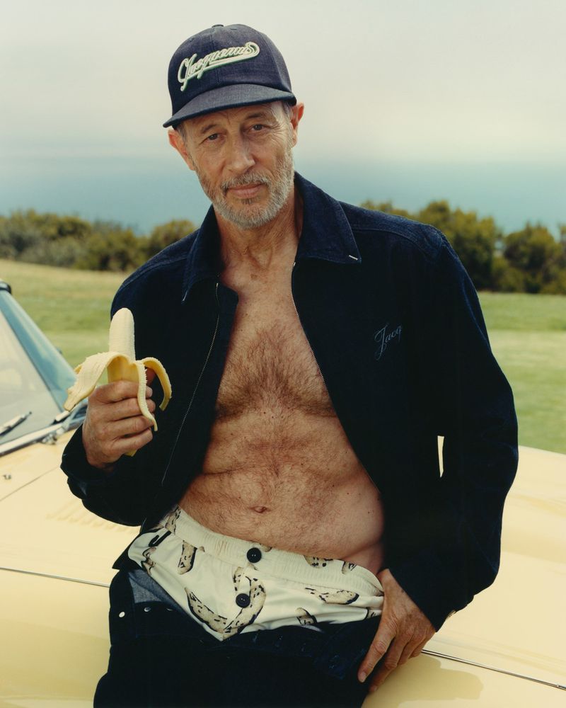 Older adult male with chest hair and baseball cap poses outdoors holding peeled banana near a yellow car on a sunny day.