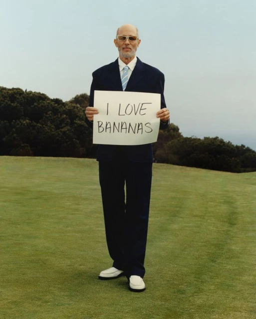 Mature man in a suit stands outdoors on a grassy field, holding a sign reading "I LOVE BANANAS", with trees and sky in background.