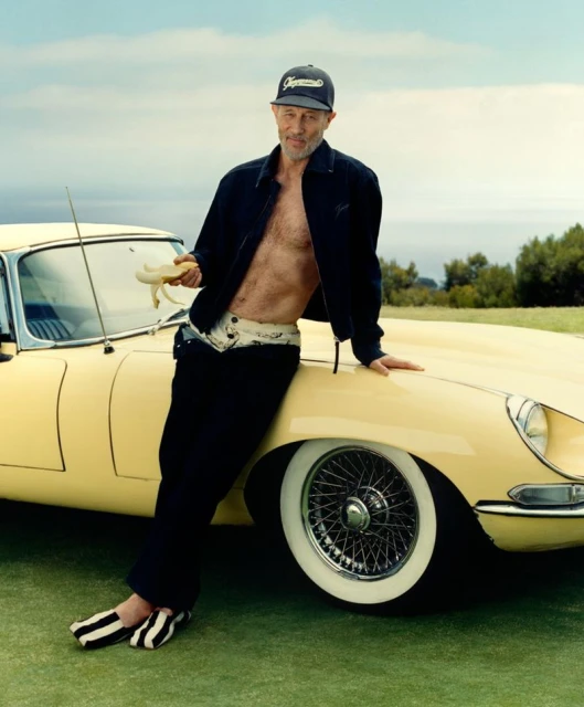 Mature man with open jacket leans on vintage yellow car with banana in hand, set against a backdrop of clear sky and distant trees.