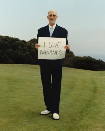 Jon Gries in a navy suit holding a sign that reads I LOVE BANANAS on a grassy outdoor course.