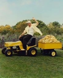 Jon Gries waving while driving a yellow lawn tractor pulling a trailer piled high with bananas outdoors