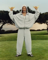 Jon Gries wearing a grey zip-up hoodie and sweatpants while holding up two bananas on a grassy course