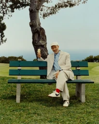 Jon Gries seated on a green park bench holding an ice cream cone, wearing a beige suit and baseball cap.