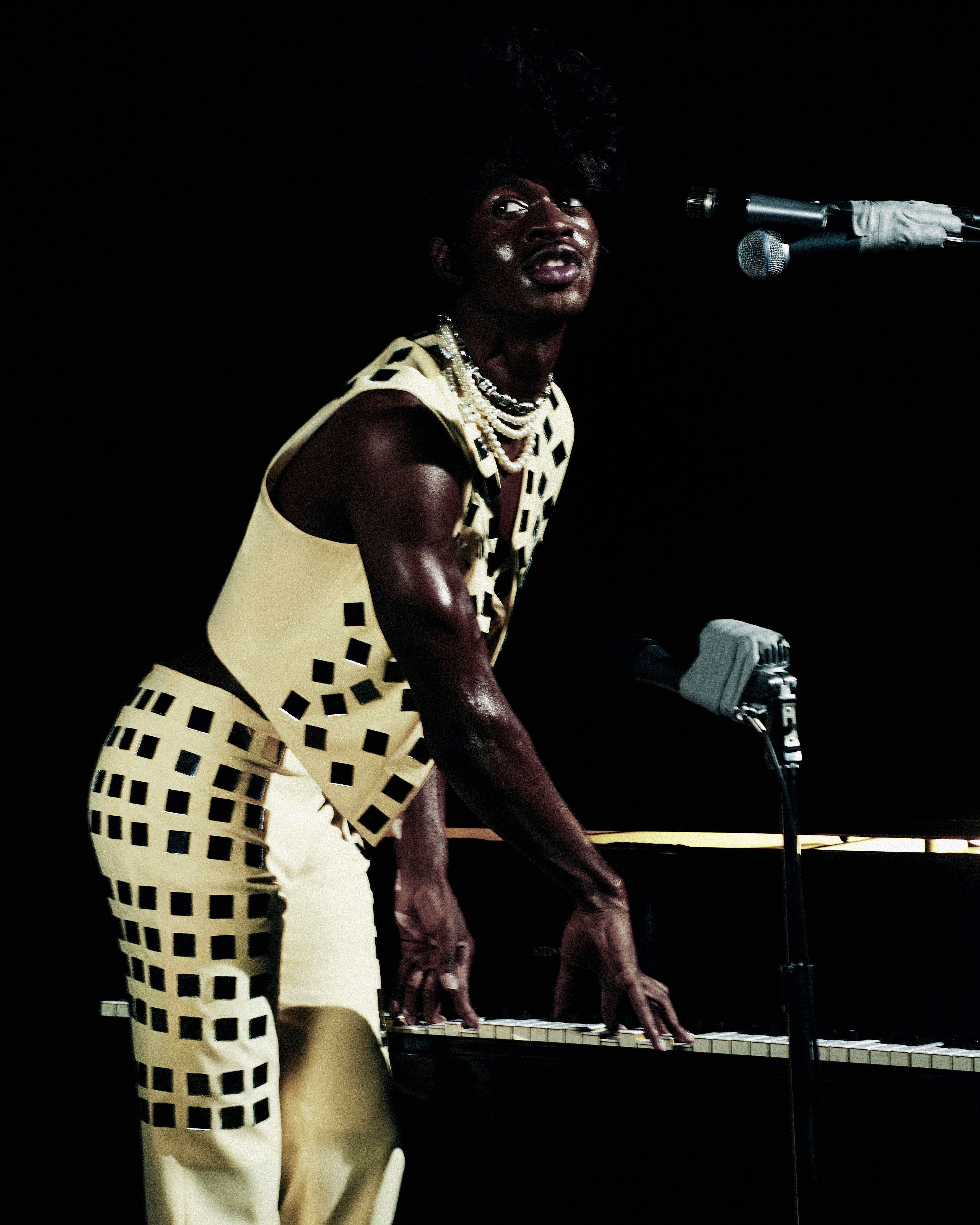 A stage view of a singer in a patterned outfit playing a piano with microphones, against a dark background, highlighting performance and artistry.