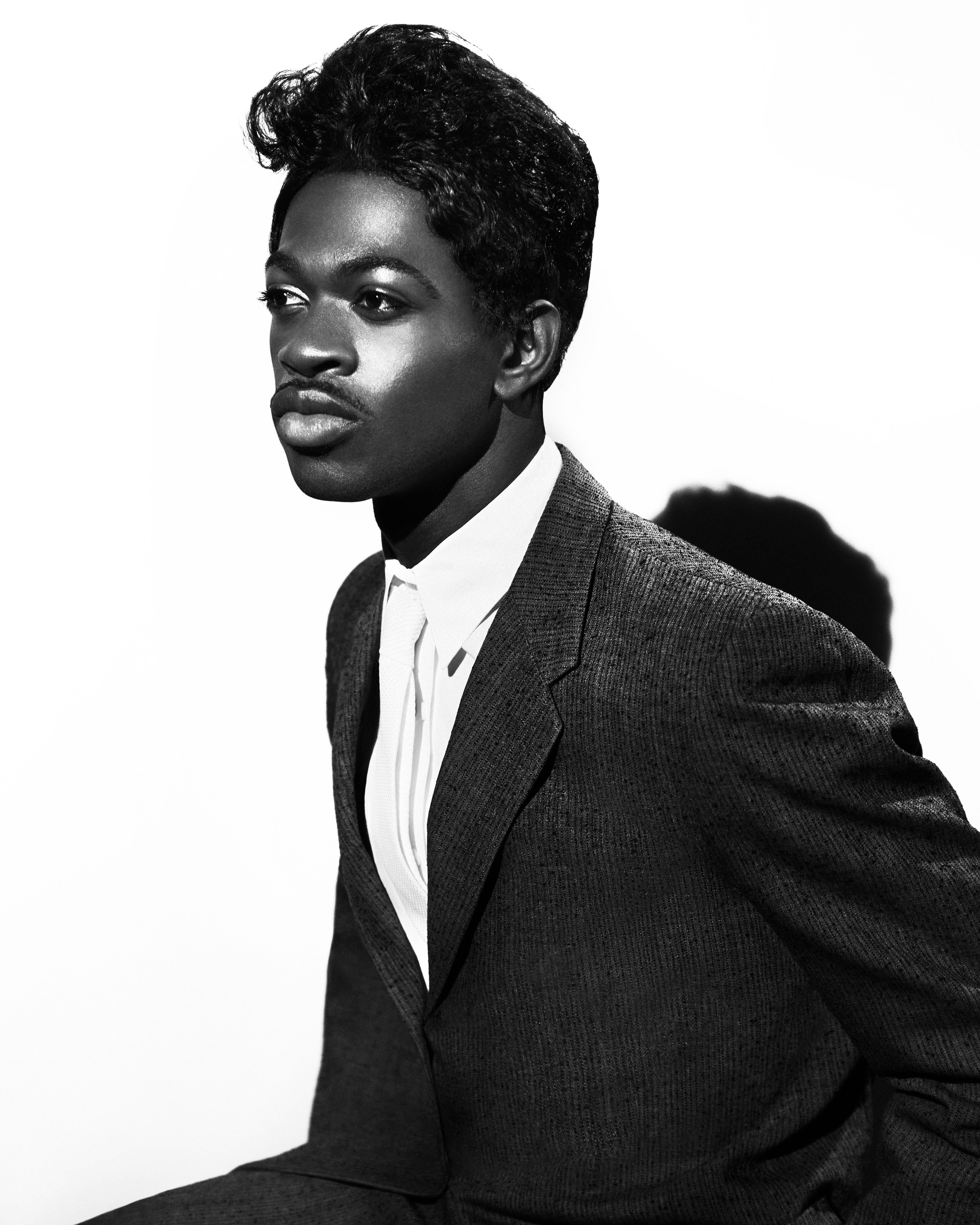 Black and white studio shot of an African American male wearing a suit, button-down shirt, and tie, looking to the side with a serious expression.