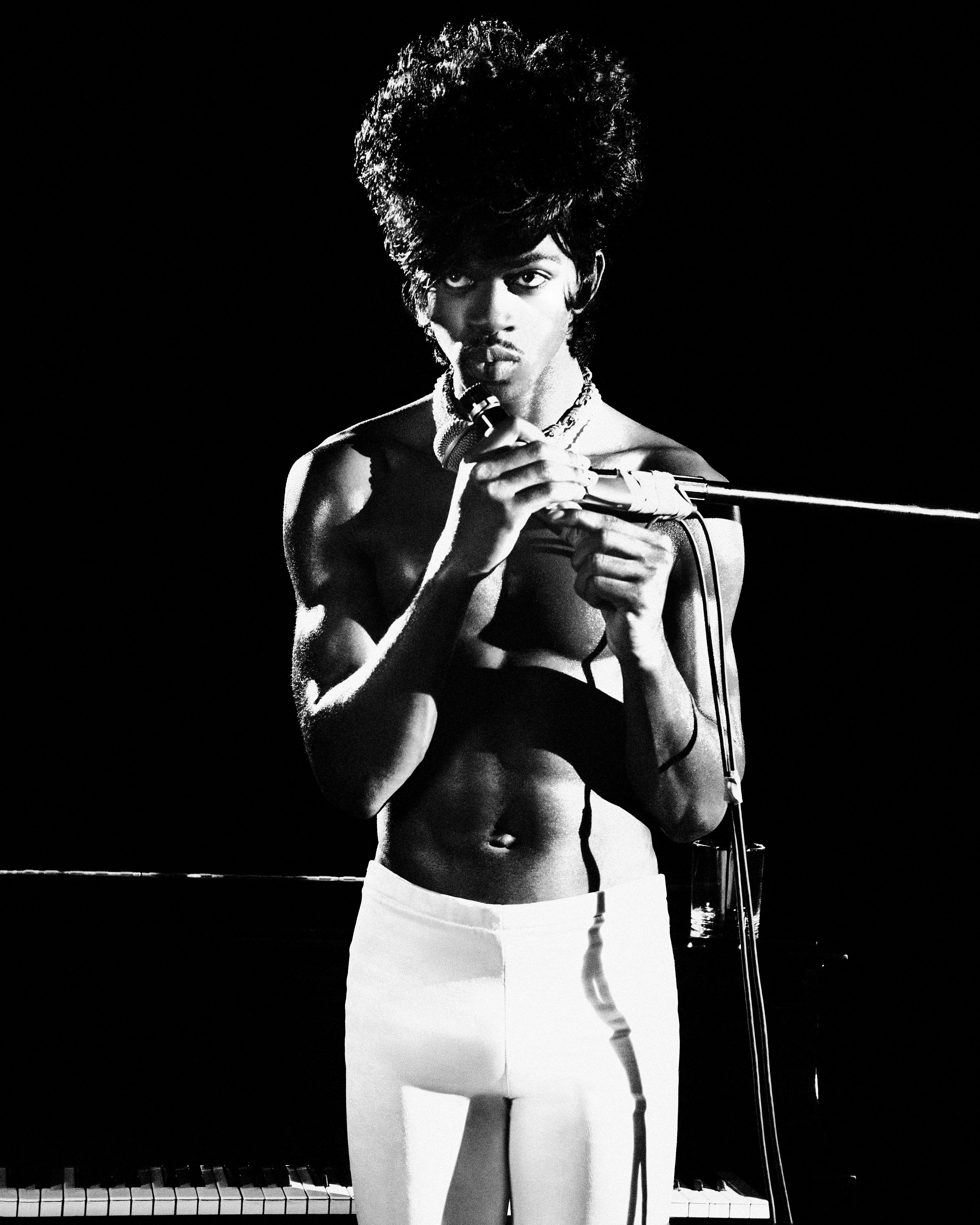 Black and white studio shot of a shirtless adult male holding what appears to be a musical instrument in front of a piano.