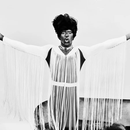Monochrome portrait of an adult with voluminous dark hair, wearing an ornate white fringed garment, dramatically posing in a photo studio.