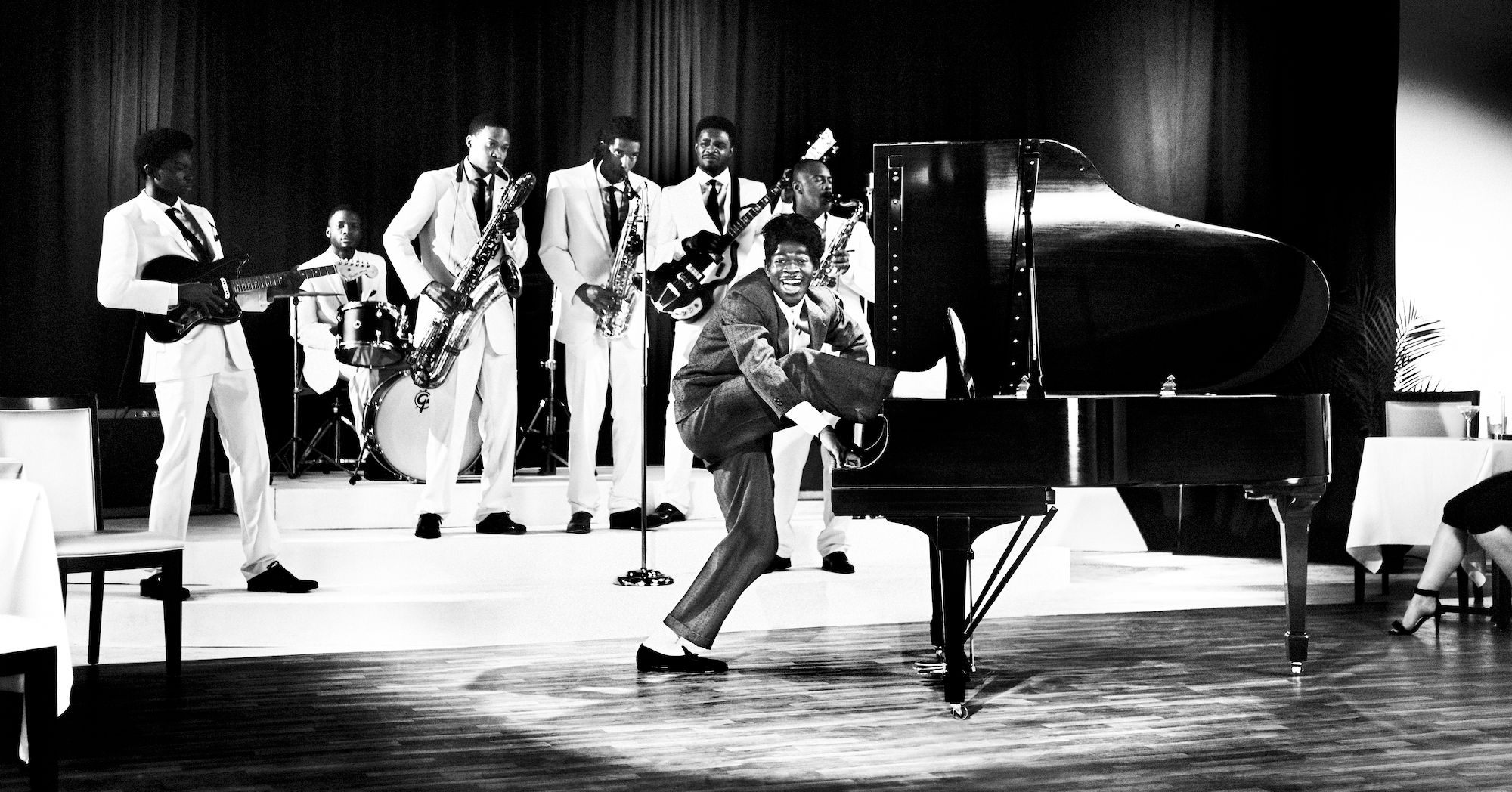 Black and white photo of a band performing with a singer standing on a piano in a room with tables and chairs.
