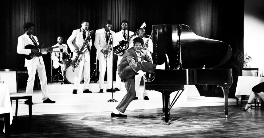 Black and white photo of a band performing with a singer standing on a piano in a room with tables and chairs.