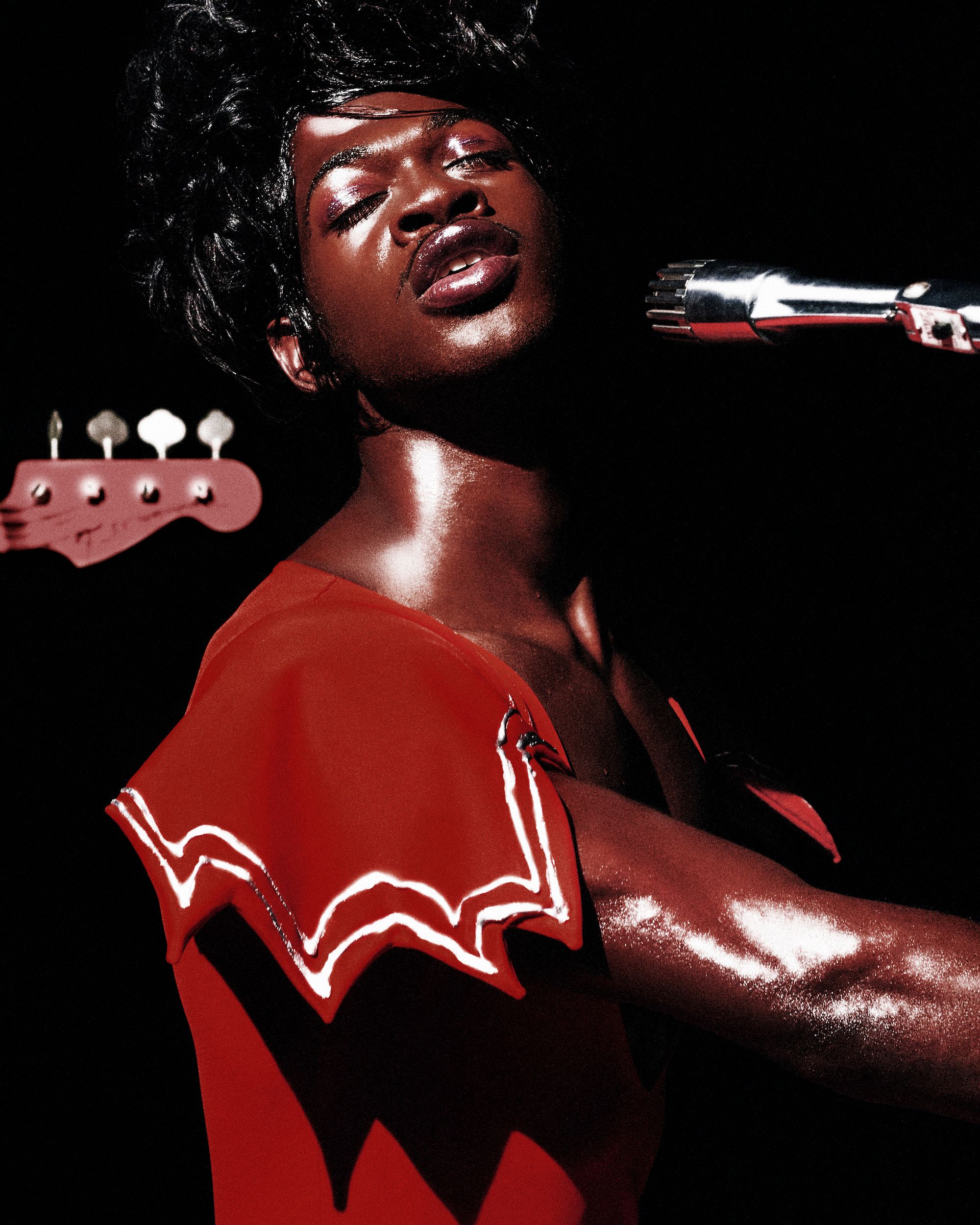 Lil Nas X poses with a microphone and bass guitar in a low-key music studio setting, wearing a red outfit and glossy makeup.