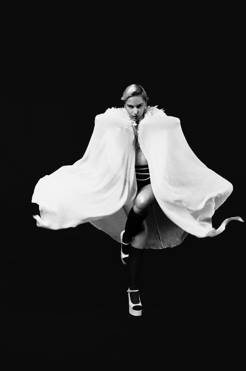 A woman in a flowing white cape poses against a black backdrop. She wears platform heels and strikes a dynamic pose.