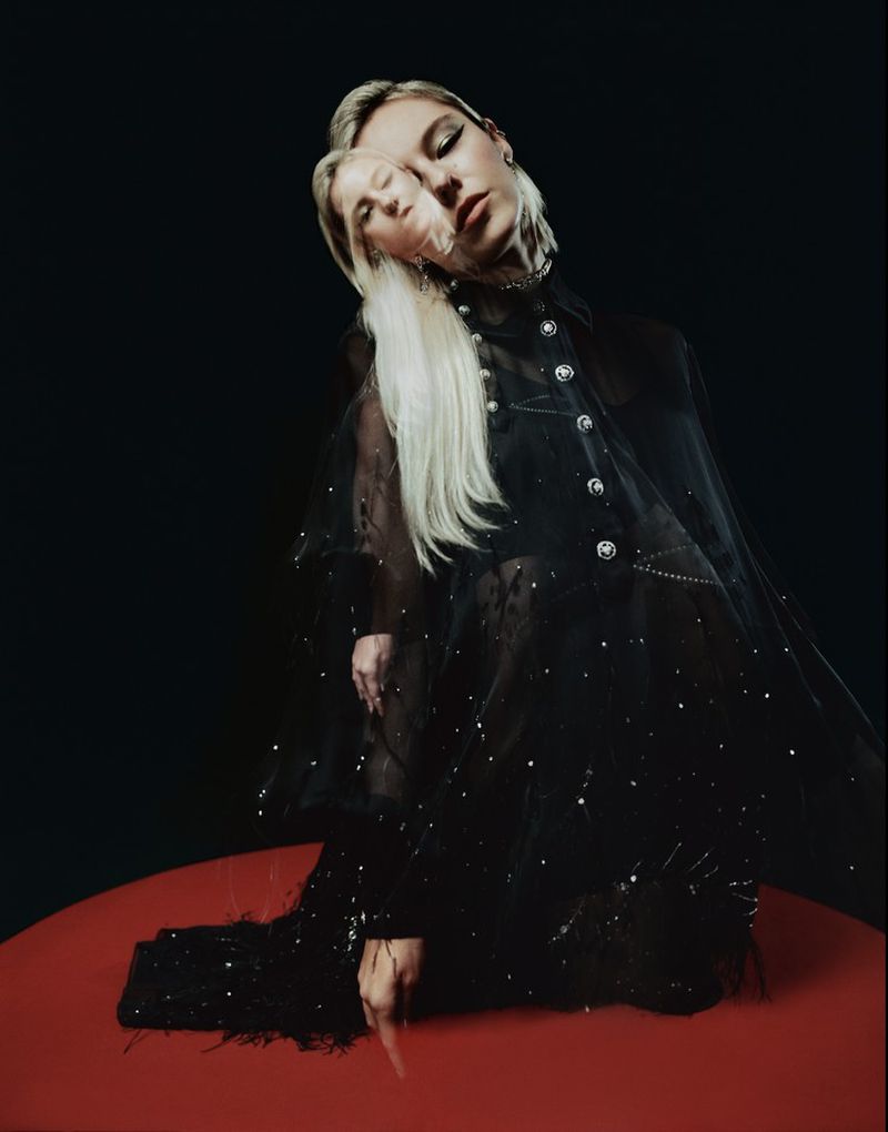 Portrait of a blonde woman wearing a black sheer embellished cape, posed against a stark dark background on a red circular platform.