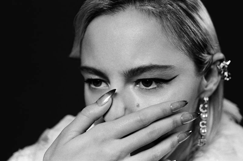 Black and white portrait of a blonde adult woman with long nails, pearl earrings, and spiky ear cuff in front of black background.