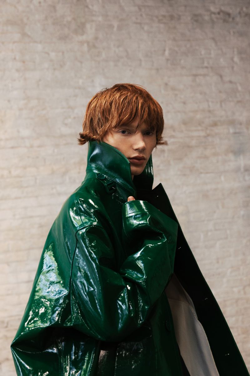 A single adult with reddish-brown hair poses in a shiny, dark green trench coat against a textured beige wall indoors.