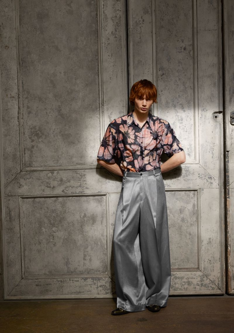 Man in floral shirt and satin pants poses against textured door. Fashion editorial shot in studio with soft lighting, stylish attire.
