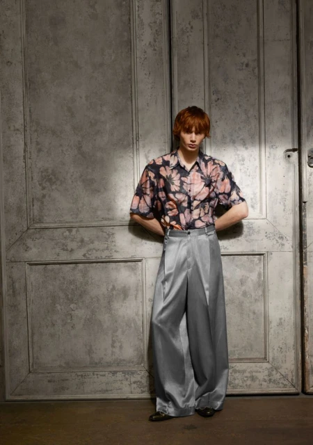 Man in floral shirt and satin pants poses against textured door. Fashion editorial shot in studio with soft lighting, stylish attire.