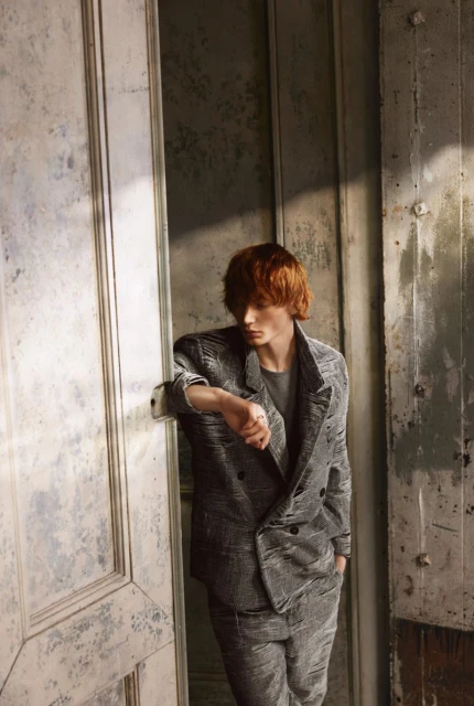 Man in a textured grey double-breasted suit leans against an aged white doorway in a naturally lit, indoor setting. Fashion editorial.