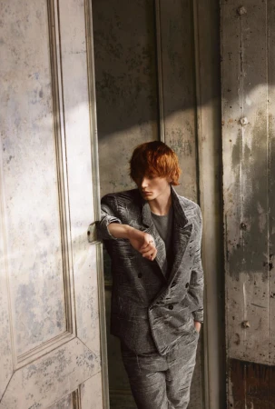 Man in a textured grey double-breasted suit leans against an aged white doorway in a naturally lit, indoor setting. Fashion editorial.